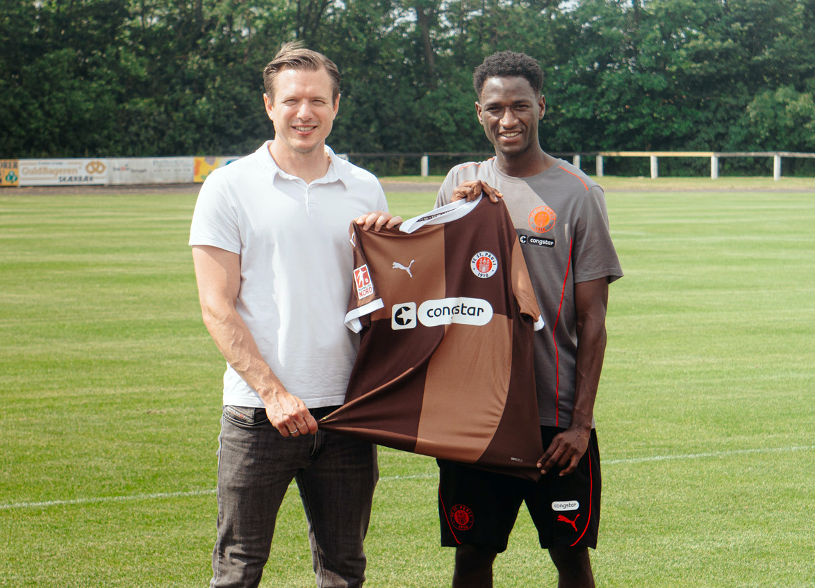 Carsten Rothenbach und Isma Baraze Adam halten gemeinsam ein Trikot des FC St. Pauli hoch, im Hintergrund ist ein Sportplatz zu erkennen.